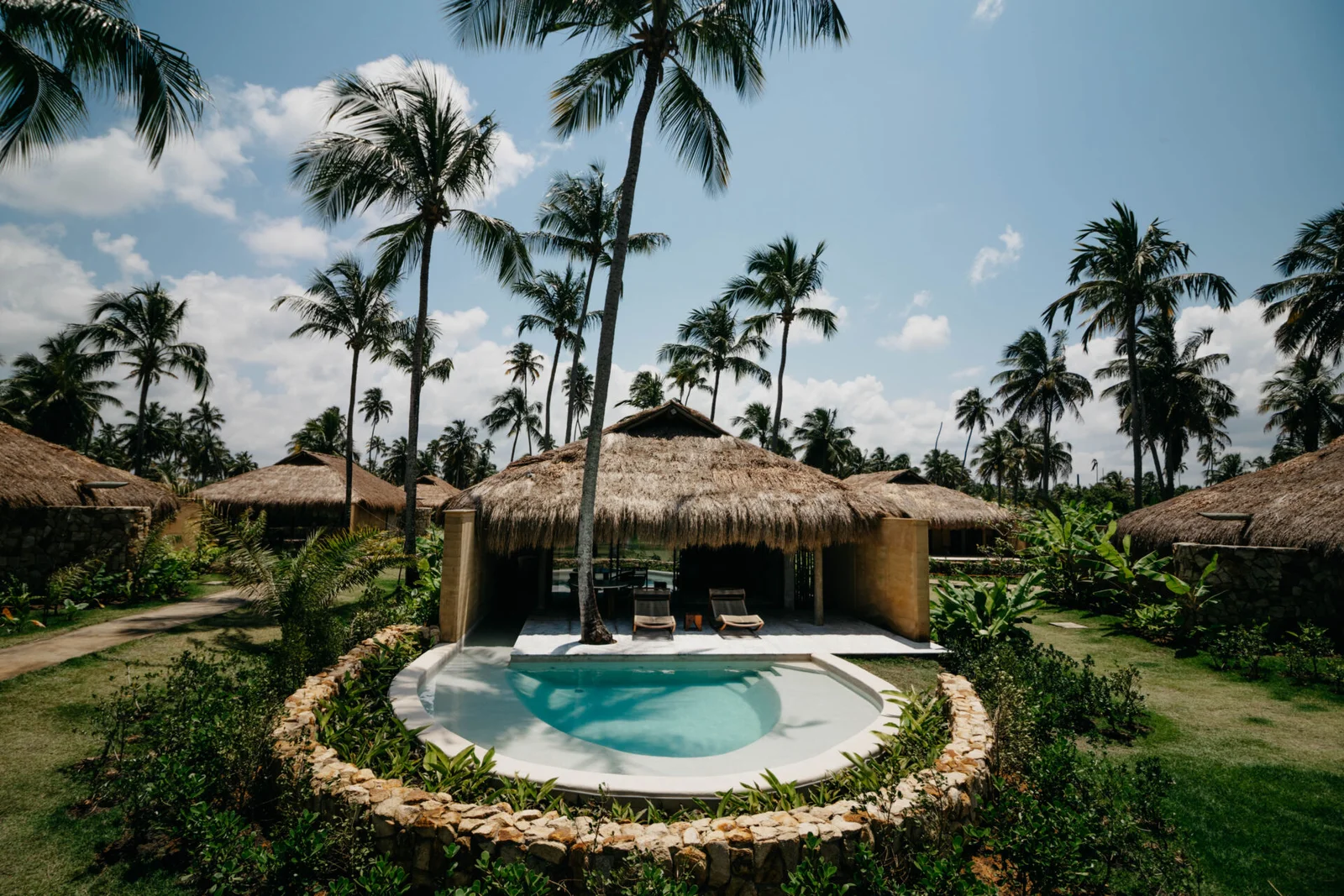 Fachada de bangalô do Primitivo Hotel & Villas, com telhado de palha, piscina e coqueiros