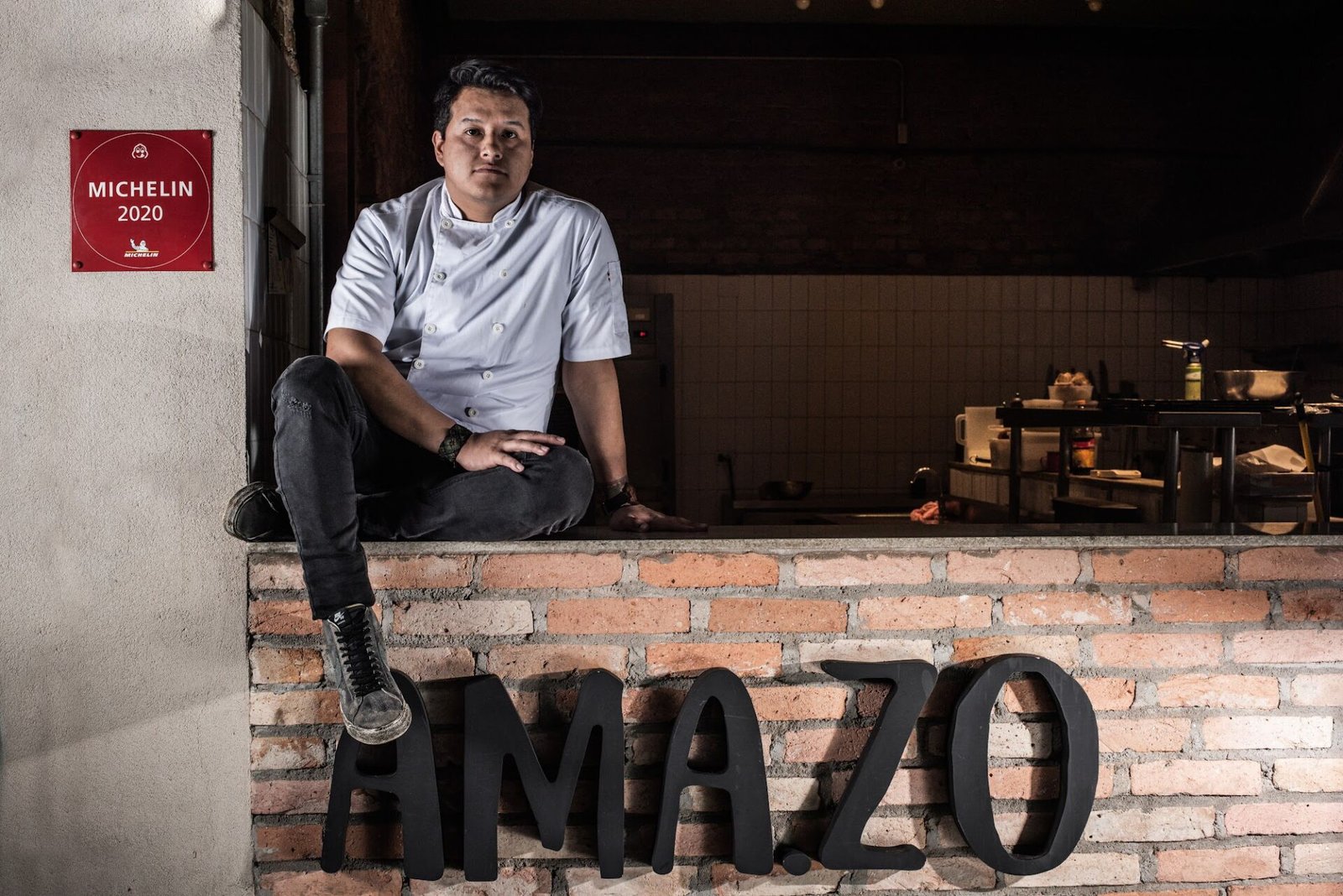 Chef Enrique Paredes sentado sobre o letreiro AMA.ZO, ao lado da placa do Guia Michelin 2020