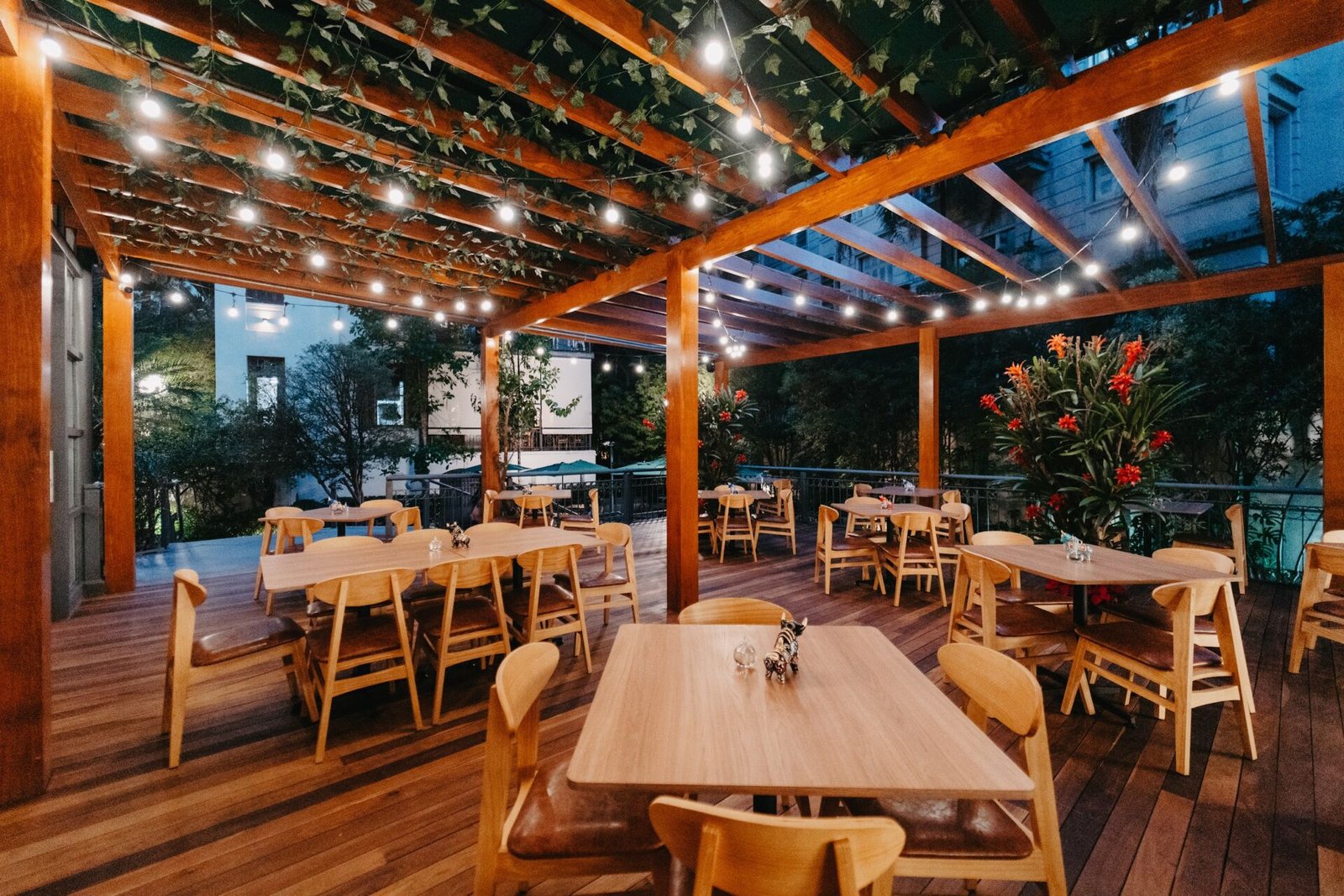 Terraço coberto do AMA.ZO no Pátio Higienópolis, com mesas de madeira, vegetação e fios de luz ao anoitecer