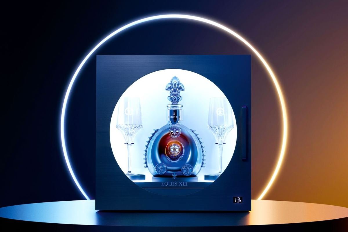 Caixa de apresentação retroiluminada da Louis XIII Black Pearl, com o decanter e duas taças em um nicho circular iluminado e um mostrador digital indicando 13 °C no canto inferior direito.