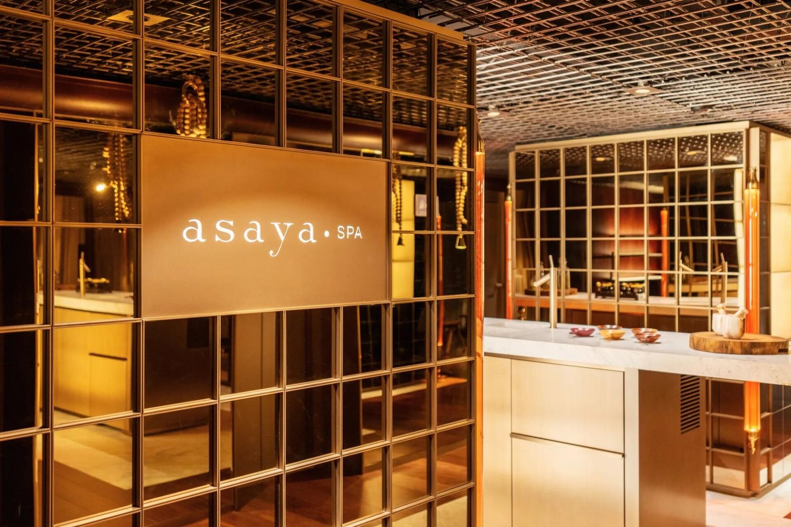 Entrada do Asaya Spa, com painel de madeira iluminado e estrutura de latão, ao lado da bancada de atendimento