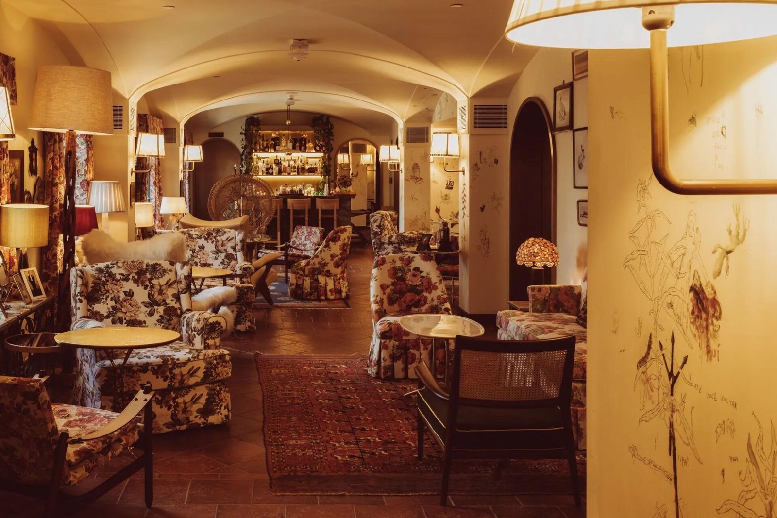 Salão abobadado do Bela Vista Bar com poltronas de estampa floral, cadeiras de palhinha, luminárias de mesa e desenhos nas paredes