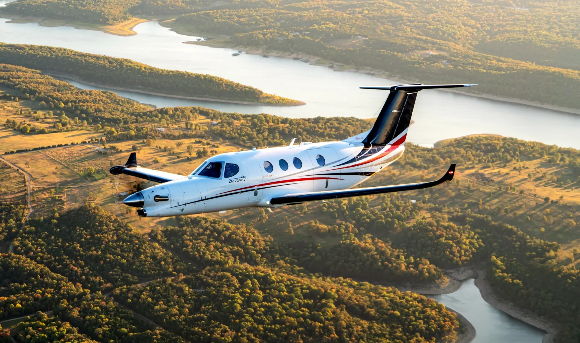 Beechcraft Denali em voo sobre vale com lago e colinas ao fim da tarde