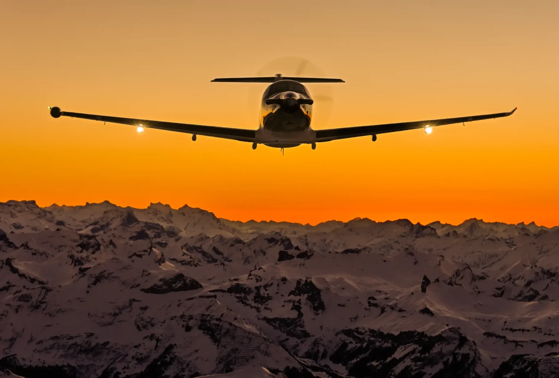 PC-12 em voo frontal contra o céu laranja do fim de tarde sobre montanhas
