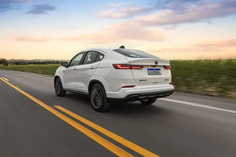 Fiat Fastback Impetus branco em três quartos traseiro, numa estrada de faixa dupla amarela entre campos, com a linha de teto descendo até a tampa do porta-malas.