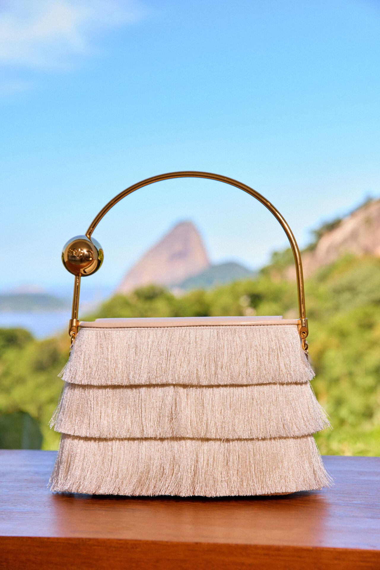 Bolsa PatBO com franjas e alça dourada com esfera, diante do Pão de Açúcar